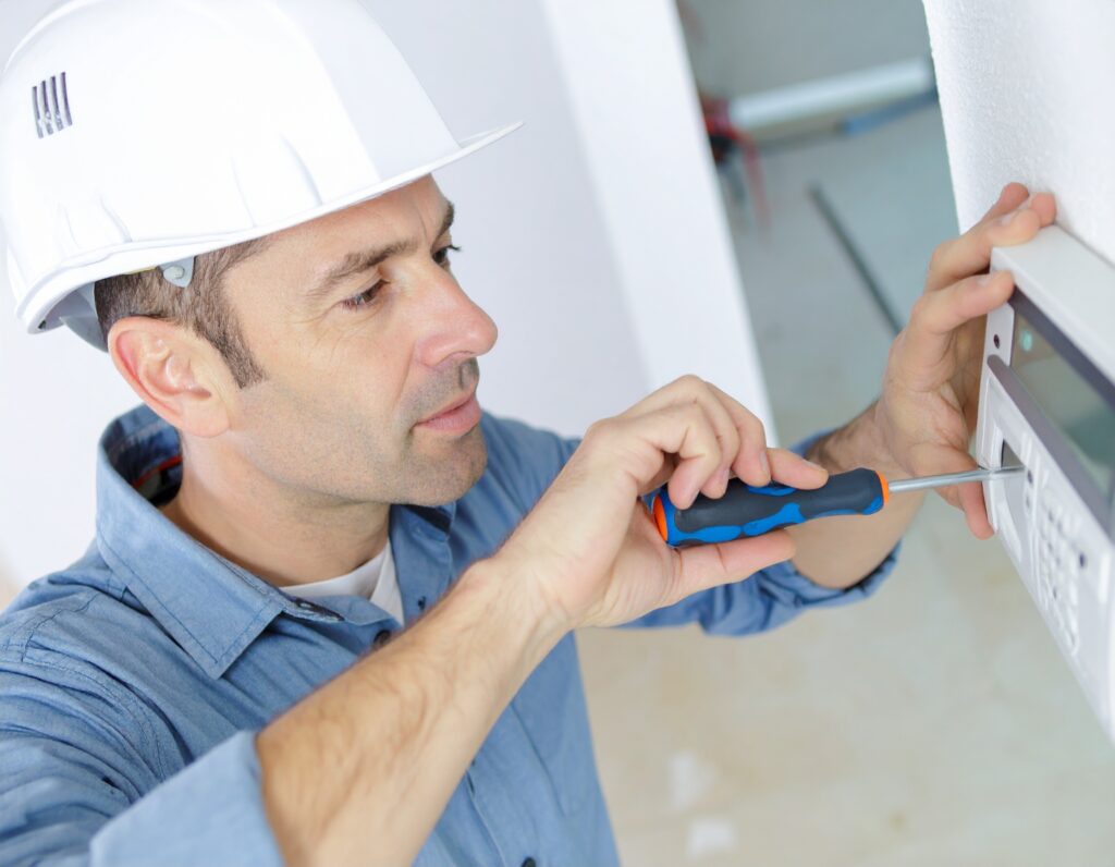 Technician installing or servicing commercial thermostat boiler control system, wearing white hard hat and using screwdriver.