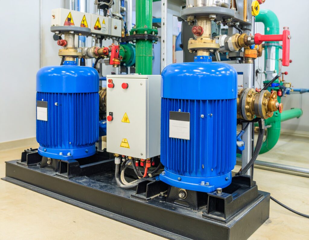 Twin water booster pump system with blue electric motors, brass fittings, and control panel in a clean mechanical room with green and red valve piping.