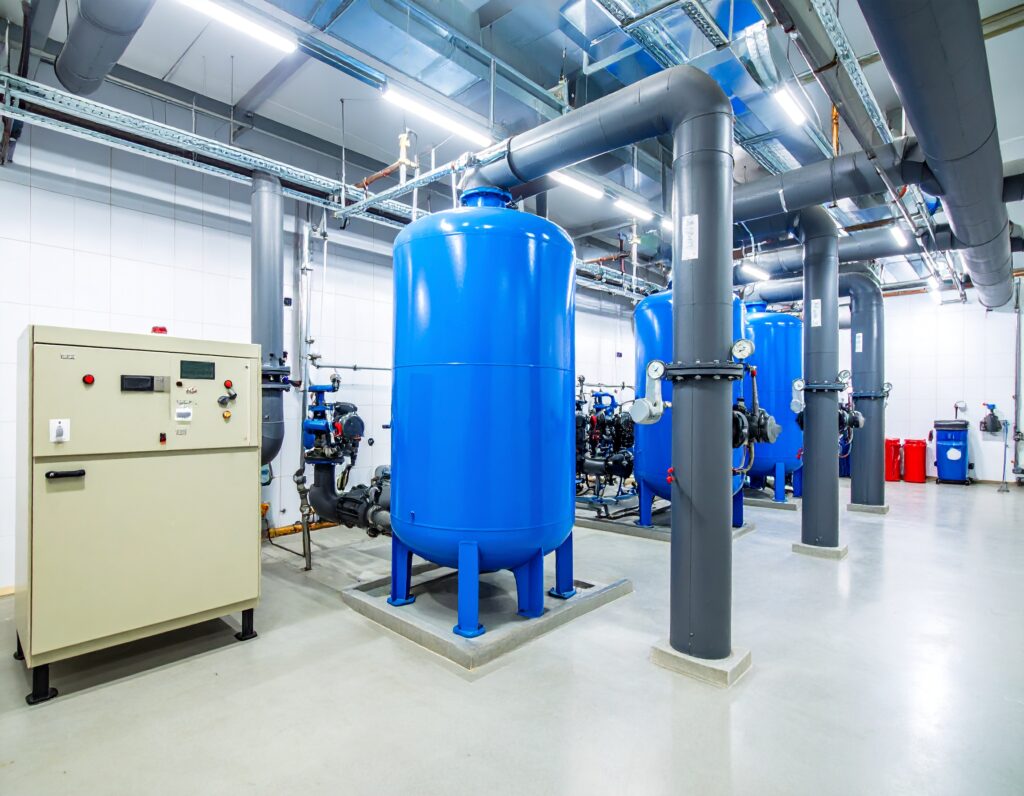 Boiler water pump replacement area featuring large blue pressure tanks, black piping systems, and a beige control panel inside a clean, modern mechanical plant room.