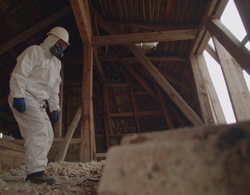 Worker in full protective gear inspecting debris in a wooden attic during an asbestos removal operation.