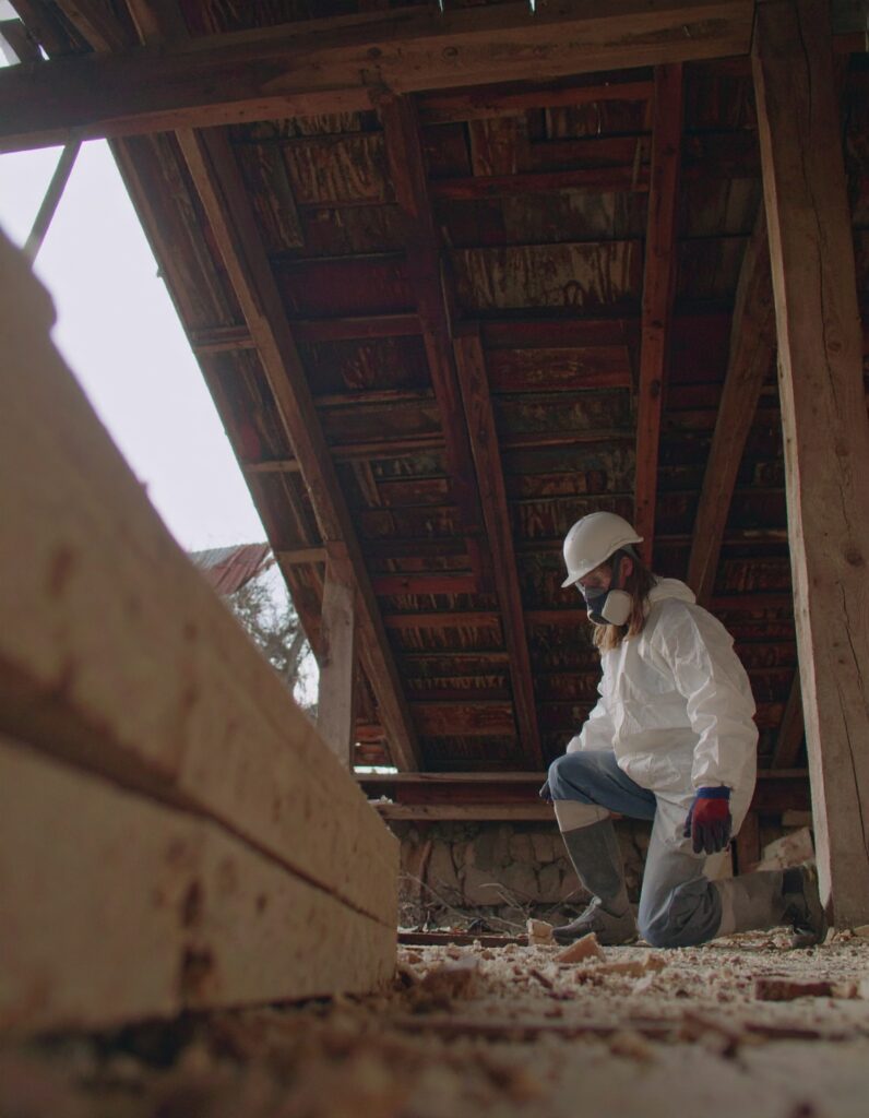 Asbestos removal technician in full protective gear kneeling in an old wooden attic during a hazardous material inspection.