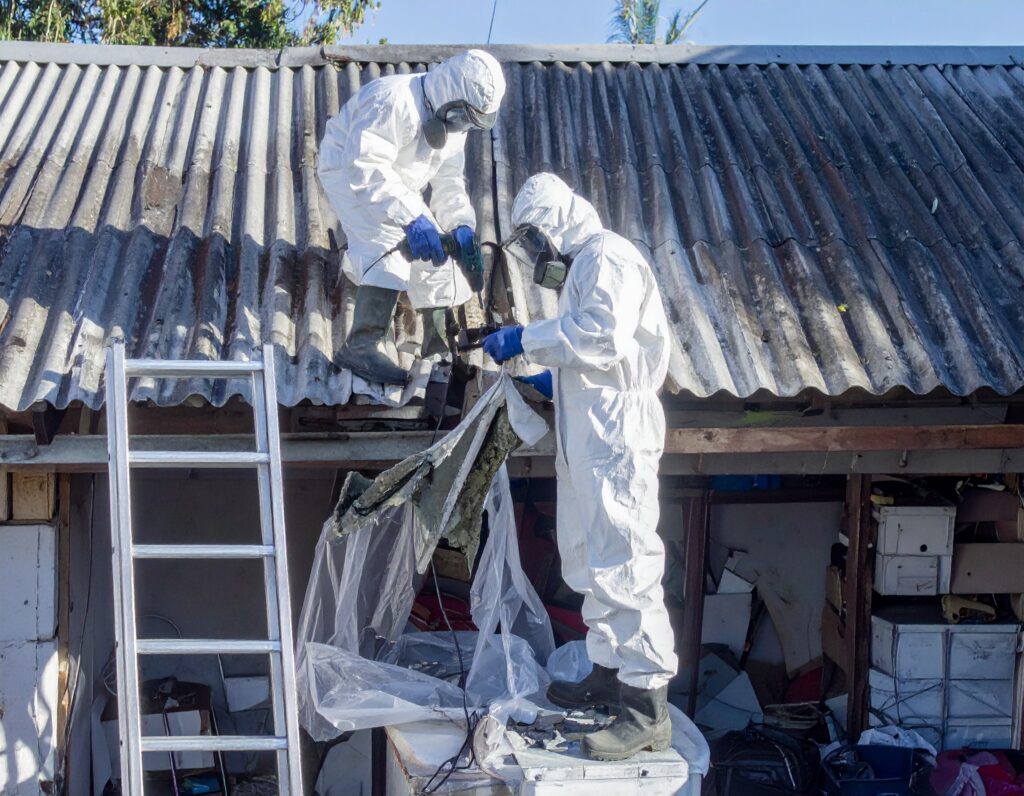Two workers in full protective suits and respirators carefully remove asbestos roofing sheets in compliance with asbestos regulations, using controlled methods to prevent fibre release.