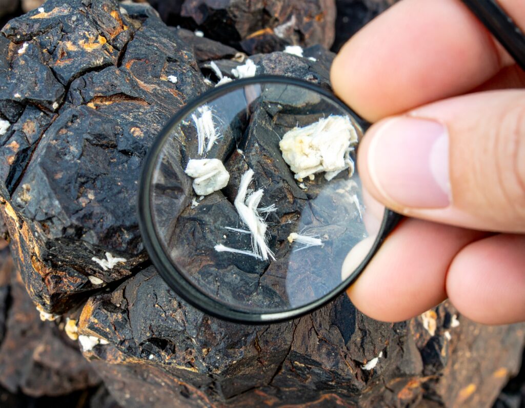 Hand holding a magnifying glass over dark rocks with visible white asbestos fibers and mineral fragments.