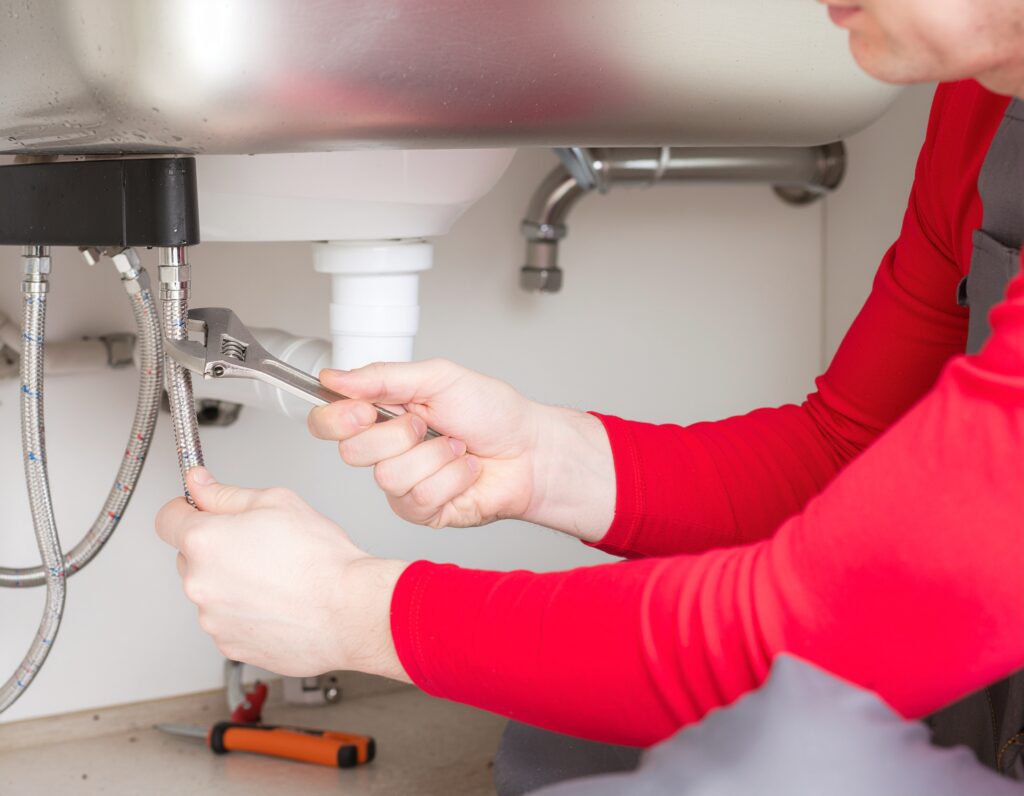 Plumber using a wrench under a sink for leak detection on flexible water supply lines.
