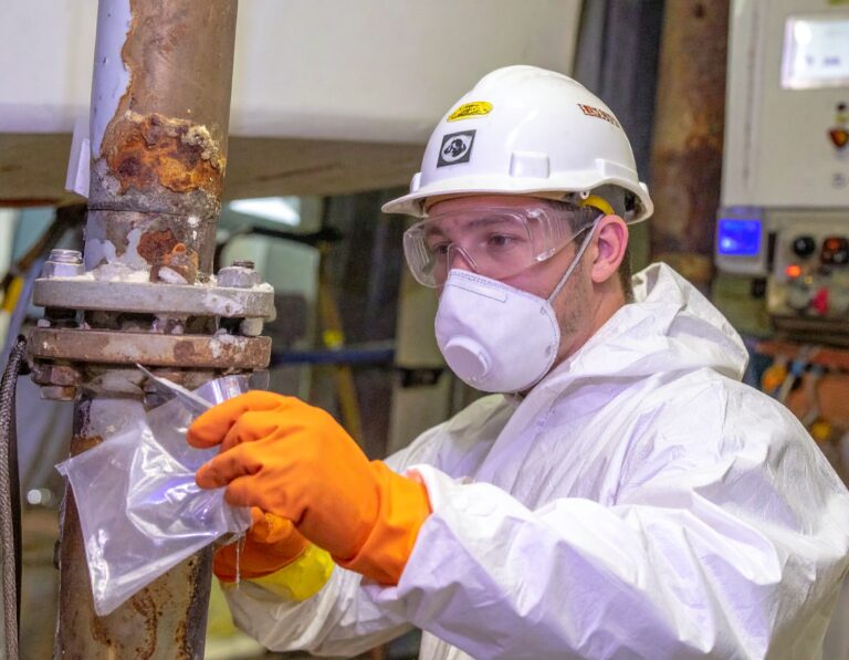 Worker in protective gear collecting pipe samples as part of asbestos removal solutions.