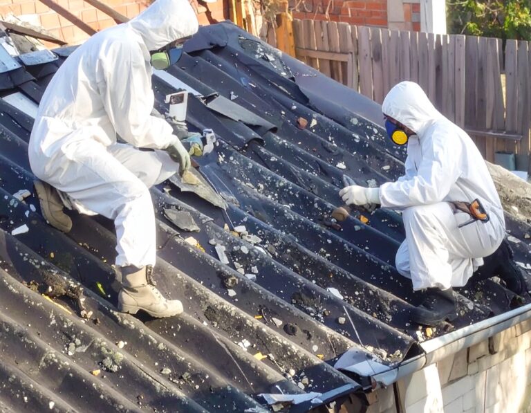 Workers in protective suits removing corrugated roofing panels during asbestos mitigation.